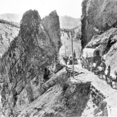 Black and white photo of a stagecoach led by a team of horses through a rocky pass above a river.