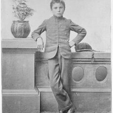 A black and white photograph of a young boy, Thomas Moore, after arriving at the Regina Indian Industrial School in Saskatchewan. He no longer has his long braids, rather a European style haircut, and he is in his school uniform which resembles a European uniform.