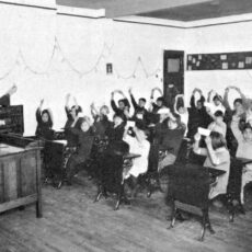 A black and white photograph of a female teacher giving a lesson to approximately 20 Aboriginal children. She is holding up a card at the front of the classroom and all the children are also holding up a card.