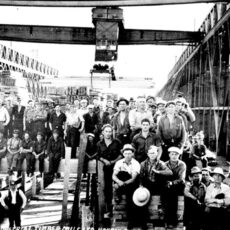 Black and white photograph of a large group of men at the Youbou Sawmill. The men are all posing for the photograph, some sitting at the front, while others are standing behind them. The photograph appears to have been taken within the sawmill as there is machinery surrounding them.