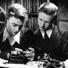 Stanley Fox and his co-director Peter Varley. Fox is holding a piece of film and both are looking at a frame, determining whether they should include it in their film. The two young men are sitting between two film reels mounted on winders.