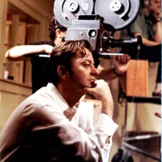 Stanley Fox crouching in the foreground with two of his crew members in the background. One of the crewmen is operating the film camera while the other is monitoring the sound recording through large headphones.