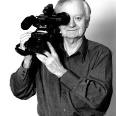 Stanley Fox poses with a large film camera propped on his right shoulder. He is wearing a collared shirt and black pants.