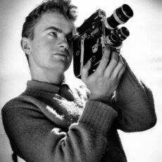 Stanley Fox posing with a Bolex movie camera. He is wearing a thick wool sweater with a collared shirt underneath. The Bolex camera is black and rectangular in shape and can be held comfortably with two hands. Fox is looking through the lens as though he is filming something.