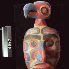 Carved wooden mask of human face with small bird head attachment on top. Mask is painted red and black.