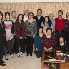 A Christmas holiday photograph of thirteen members of the Loy Sing Guen family in 2013, all casually dressed and smiling broadly. In the foreground is a table with a box of chocolates and other holiday treats on it.