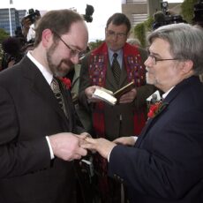 Two men exchanging wedding rings and vows. A United Church minister stands between them holding a book.