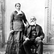 A black and white studio portrait of woman standing and her husband seated. They are wearing formal clothing.
