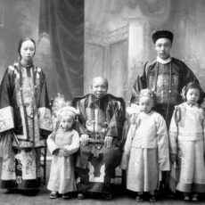 A studio portrait of Lee Mong Kow’s family in traditional clothing. In the middle sits an older woman, with three young children around her. Behind them stand a young woman and a young man.