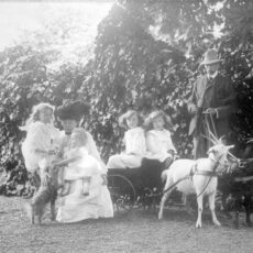 A black and white photograph of the Burns family shows a father holding the reins of two goats hitched to a buggy with two children in it. The mother, two other children and a dog are next to them.