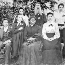 A black and white photograph of a family sitting outside. In the centre sits an older woman in a dark dress. Behind her stands an older man with a bushy beard. Around them are five young people, including teenage boy with a dog at his side, two young girls and two young women.