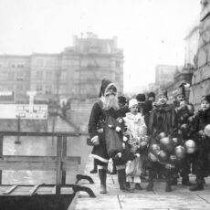 A black and white photograph showing Santa Claus standing on Victoria’s Inner Harbour causeway with a group of young boys dressed as clowns standing next to him.
