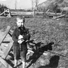 A black and white photograph showing a little boy standing barefoot in a field holding a black kitten.