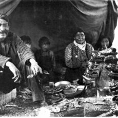 A black and white photograph showing a First Nations family sitting in a tent smoking salmon heads on sticks over a small fire.