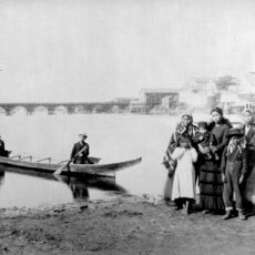 A First Nations family. Four women and four children stand on the muddy shore, while two men sit in a wooden canoe in the harbor. A bridge and some wooden buildings are in the background.