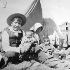 A black and white photograph of a First Nations man seated on the ground holding a baby. A woman sits next to him with a toddler on her lap.