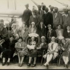 Image of people on a ship's deck in 1931. Front row are women sitting down. Middle row are women standing. Back row are men standing in suites. Person in front row has life preserver around her head.