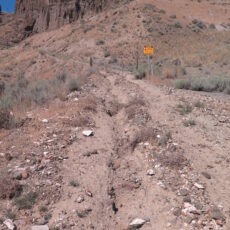 A rugged, unpaved road up a dry mountain covered in rocky outcroppings. A sign by the side of the road says “Danger! Rockfall Risk Area” and an old metal gate blocks the road.