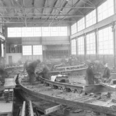 Workers construct sections of railway tracks. Steel and boards in piles on factory floor.