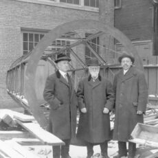 Three men wearing heavy coats in front of telescope tube. There is snow on the ground.