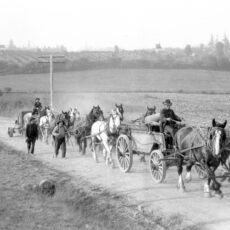 Two carts, ten horses and five men haul equipment along a dirt road. Farmland and rolling hills in the background.