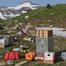 Camping and field work equipment are spread out on the ground in an alpine area. There is a snowy mountain in the distance.