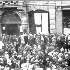 A large crowd stands waiting on the street in front of buildings on Government Street in Victoria BC. Long lists of paper are posted on the windows of the building. The crowd is mostly men wearing hats and suits. A few men in soldier’s uniforms are visible as are a few women.