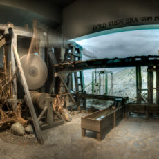 A giant wooden moving wheel and troughs in front of a painted backdrop showing dry hills.