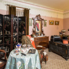 Image of a bedroom from the 1800s that is covered in pink wallpaper and includes a standing clock, a sitting table, and a wooden bed.