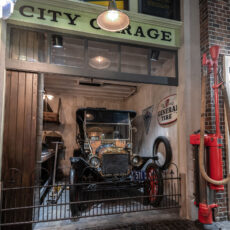 A black 1900’s vehicle sits in a garage that is gated off from the wooden cobblestones streets.