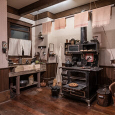 A 1890s kitchen with a wooden sink and iron stove. Area is full of cooking tools such as a waffle iron, kettle, and plates.