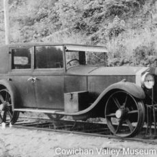 An old 1930s car with unusual metal wheels parked on train tracks, with shrubbery in the background.