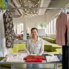 A woman sits behind a table smiling. On the table are garments that are folded and displayed including knitted sweaters and embroidered pillowcases. In front of this table is a rack of hanging shirts.
