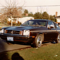 A brown car, a Chevrolet Monza, sits parked in a driveway on a residential street in 1983.