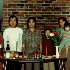 A family of recent Vietnamese migrants pose for a photo standing behind a dining table in a residential home. The table is set with candles, incense, bowls of citrus fruit and a roasted chicken. Behind the table stands a young new mother holding her one-month-old baby, three young adult men, a nine-year-old boy and another toddler being held by one of the young men.