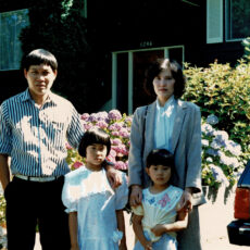 A Vietnamese family composed of an adult man, an adult woman and two young girls stand in the driveway of a suburban home in Victoria, BC, beside a blue Ford Escort station wagon. There are hydrangea bushes full of flowers behind them with a dark brown single detached home in the background.