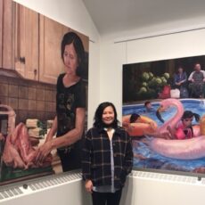 A woman stands between two colourful paintings. On the left is a painting of a woman cutting up chicken. On the right are children playing in a backyard pool. Parents watch from the side.