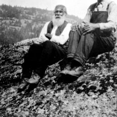 Two men sitting outside on a hilltop. There is another hill in the distance. The man on the left has a white beard and moustache, long pants, white shirt and dark vest. The man on the right is wearing overalls.