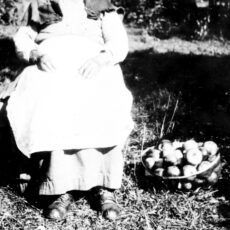 A woman sitting on a chair outside. She is wearing a hat and a long dress. There is a container of apples beside her on the ground.