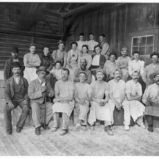 Twenty-four people, workers at a factory, are seated and looking at the camera. Most are in work clothes. The owner is seated, second from the left in the front row, leaning on a cane.