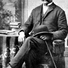 Portrait of a man with a moustache sitting in a chair, wearing a suit, holding a cane and hat. Books on a table are in the background.