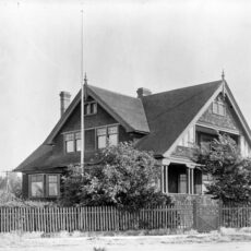 Black and white photograph of a house with many windows, a picket fence and trees outside of it.