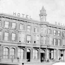 Black and white photograph of a three story hotel with many window, a turret at top in the centre and people at street level in front of the building.
