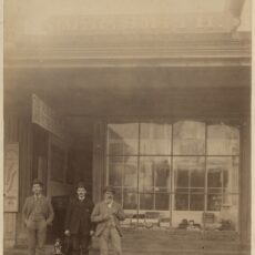 Three men in suits stand on a wooden sidewalk outside of a business. The man in the middle has a dog on a leash. A storefront window is behind them.